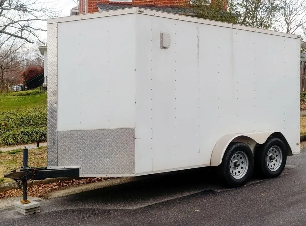 A white enclosed utility trailer with two axles and a silver diamond-plate panel on the front is parked on the side of a residential street